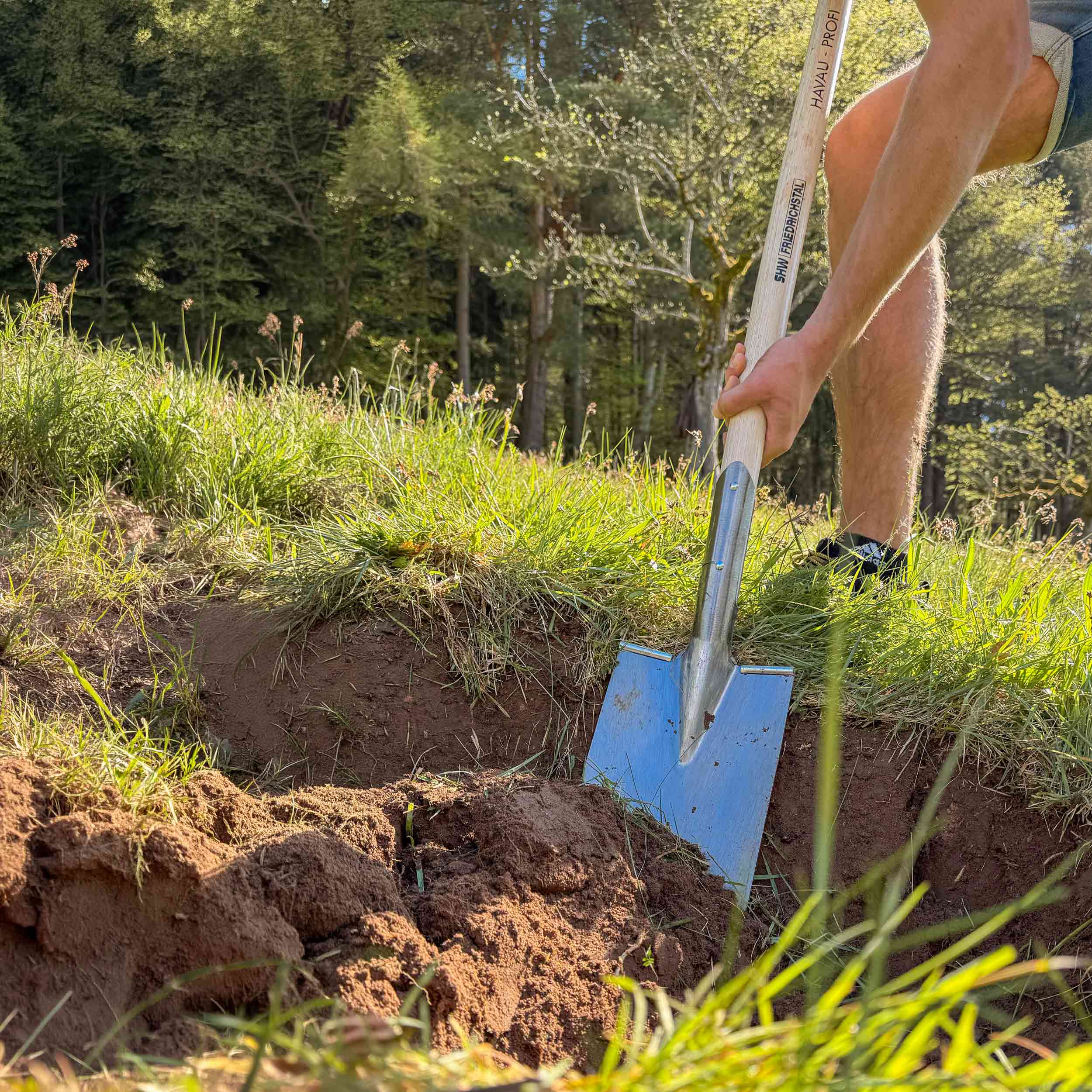 Person arbeitet mit Edelstahl-Drainagespaten mit langem Holzstiel beim Ausheben eines Grabens im Garten – robuster Grabespaten für Gartenbau und Landschaftspflege